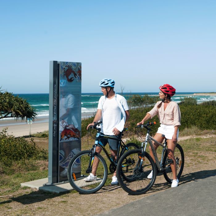 Image of a couple enjoying a bike ride on the shared coastal pathway trail in Ballina featuring Aboriginal signage acknowledging local cultural heritage in the North Coast region