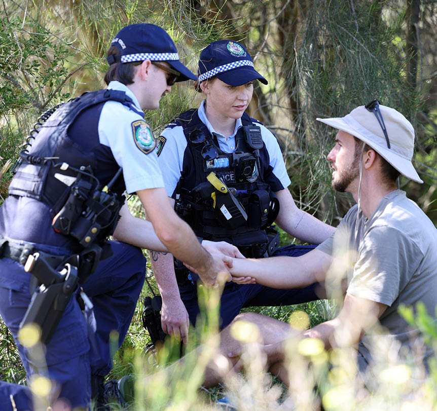 Two police officers helping a person sitting on the ground in a bush area