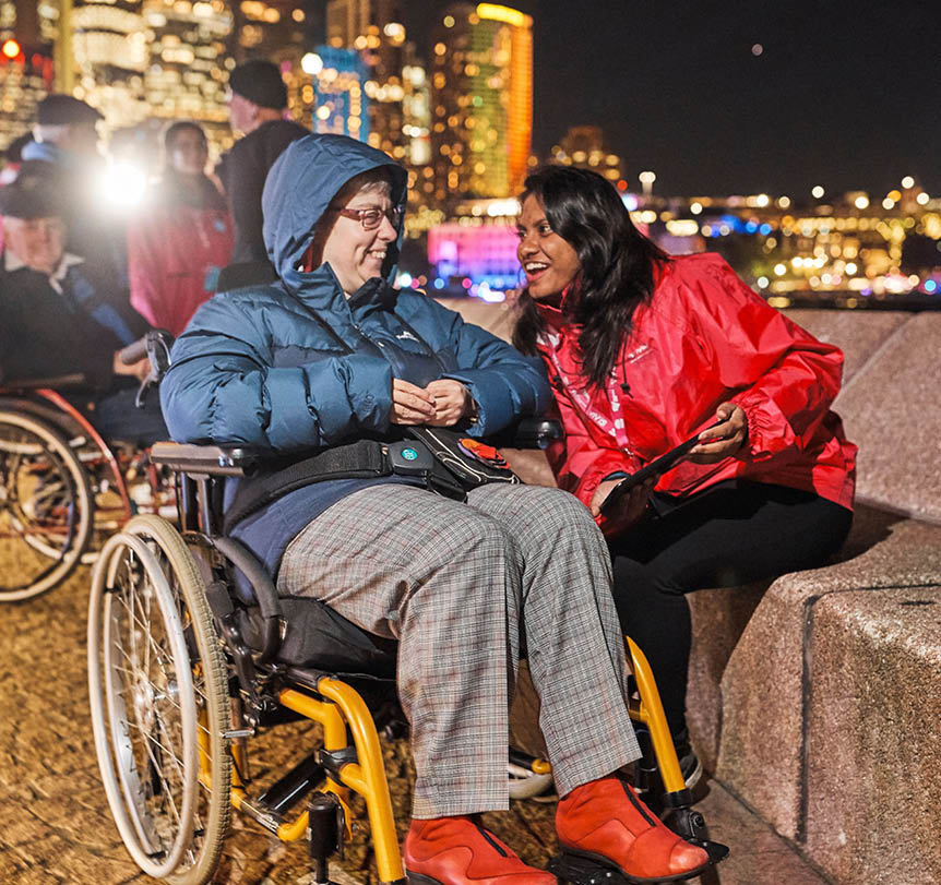 A person in a wheelchair talking to a person holding a digital tablet outside at night with city lights in the background