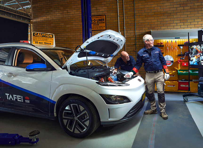 Two people in a workshop standing next to a car with the bonnet open.