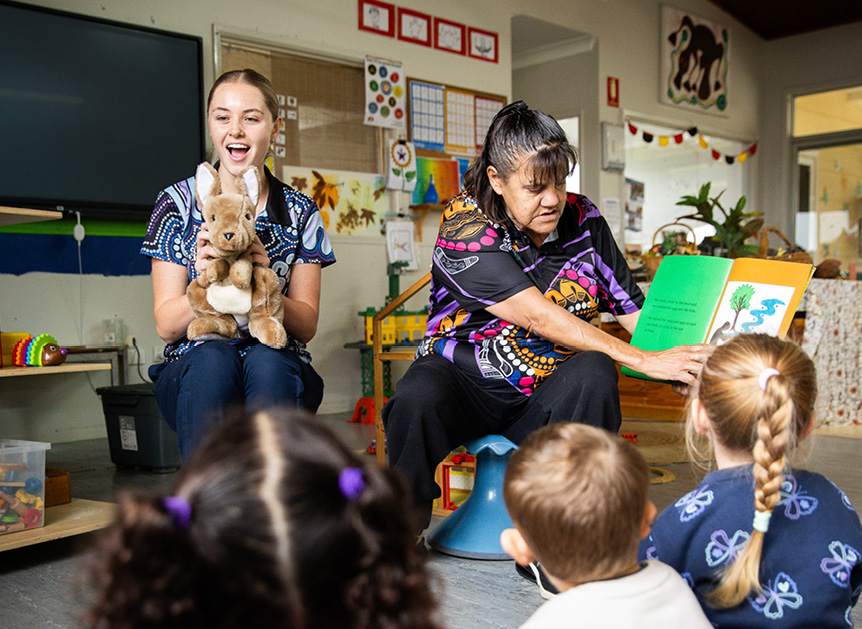 Two preschool teachers with a plush kangaroo reading to a group of children sitting on the floor.