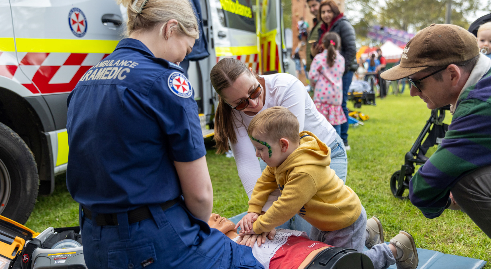 A child practicing CPR on a dummy with their parents and paramedics at a festival