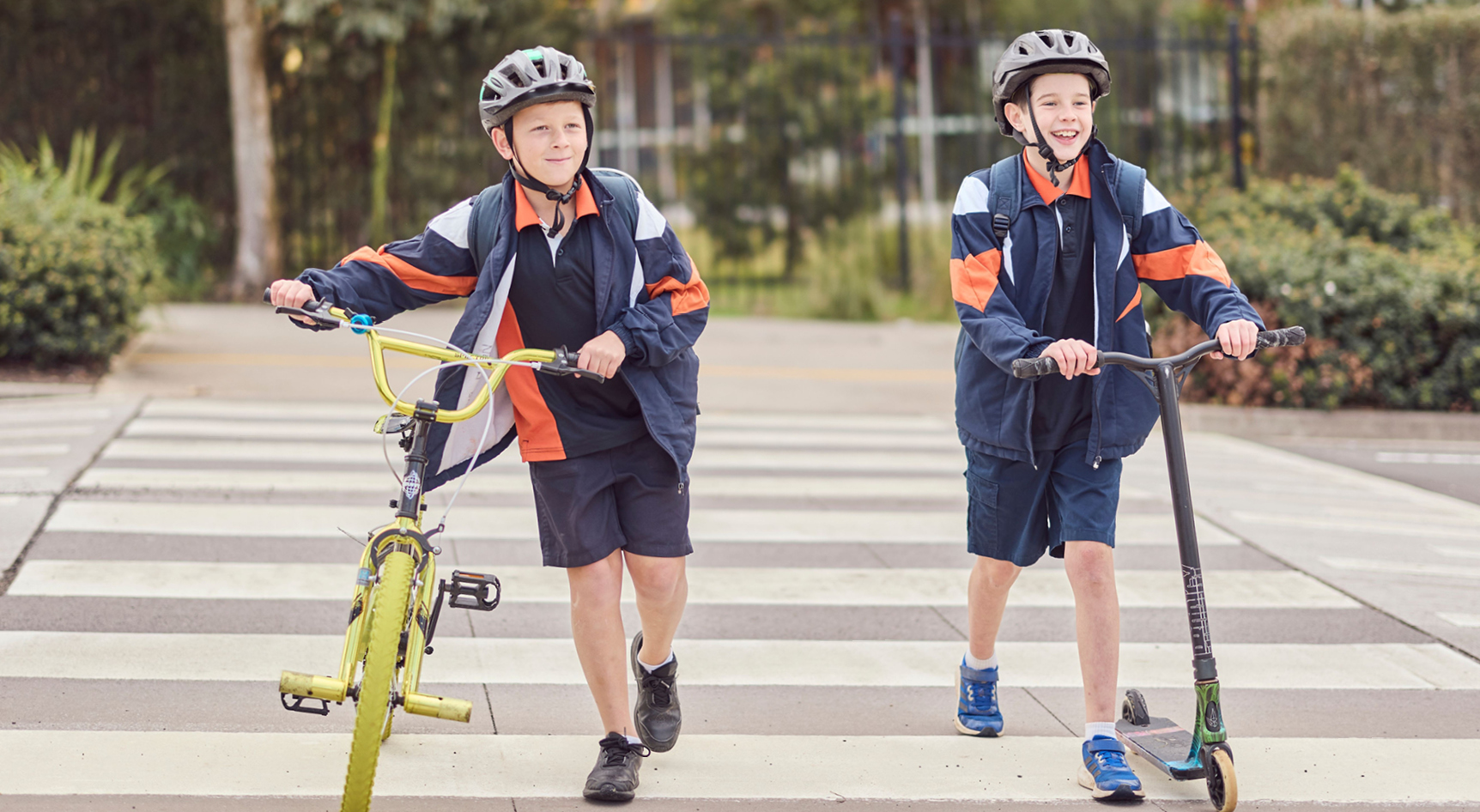 Two children wearing helmets walking across a school crossing with a bike and a scooter